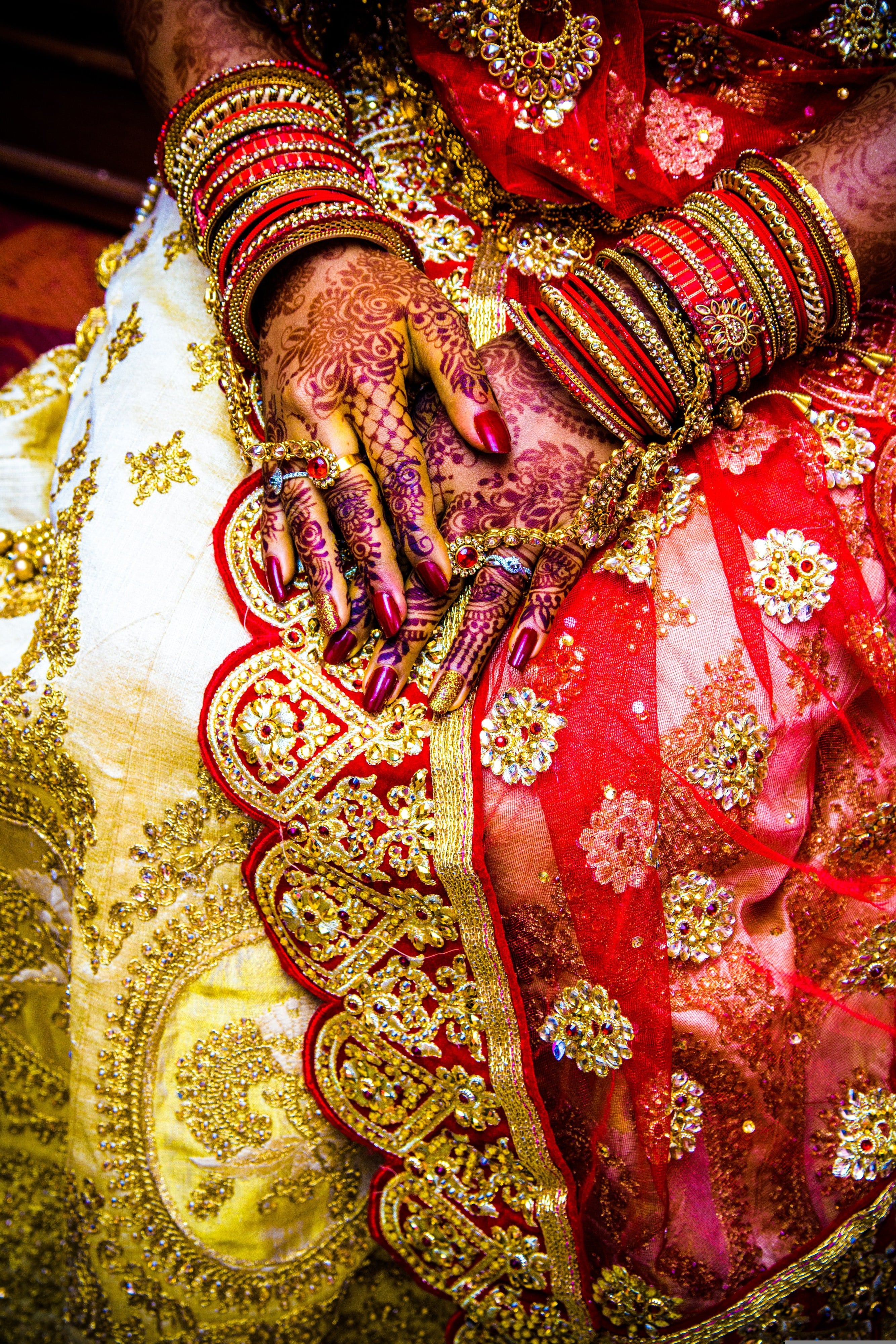 A bride’s hands with mehendi and red bangles, resting on her embroidered lehenga